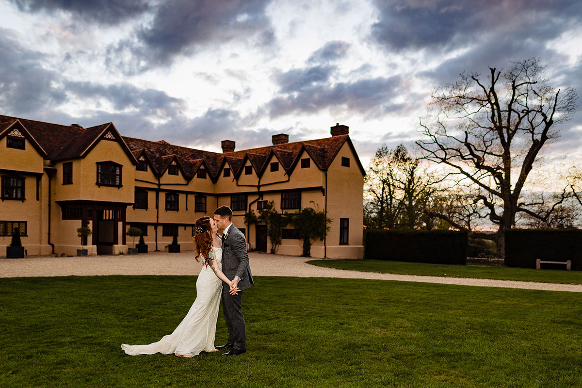 A bride & groom kiss romantically while holding hands in front of the main house at Upton Court