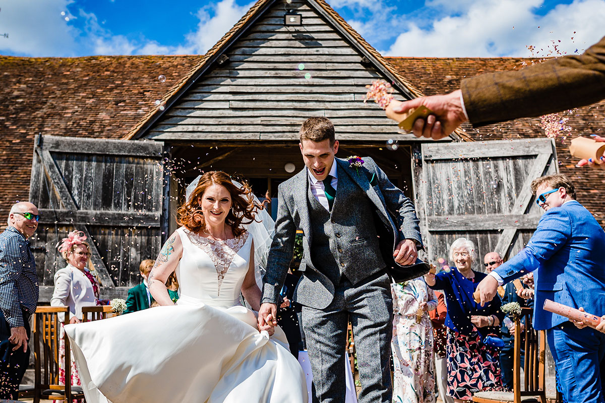 Groom wearing a grey tweed suit holds hands with a bride with red hair wearing an ivory dress. They are jumping a broom in front of the barn at Upton Court wedding venue