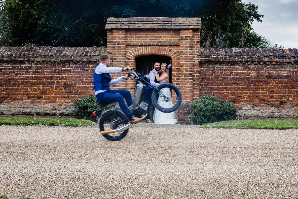 Colour photograph of a man riding a motorbike on one wheel while passing a nride & groom. bride & groom are embracing under an arch in a red brick wall.