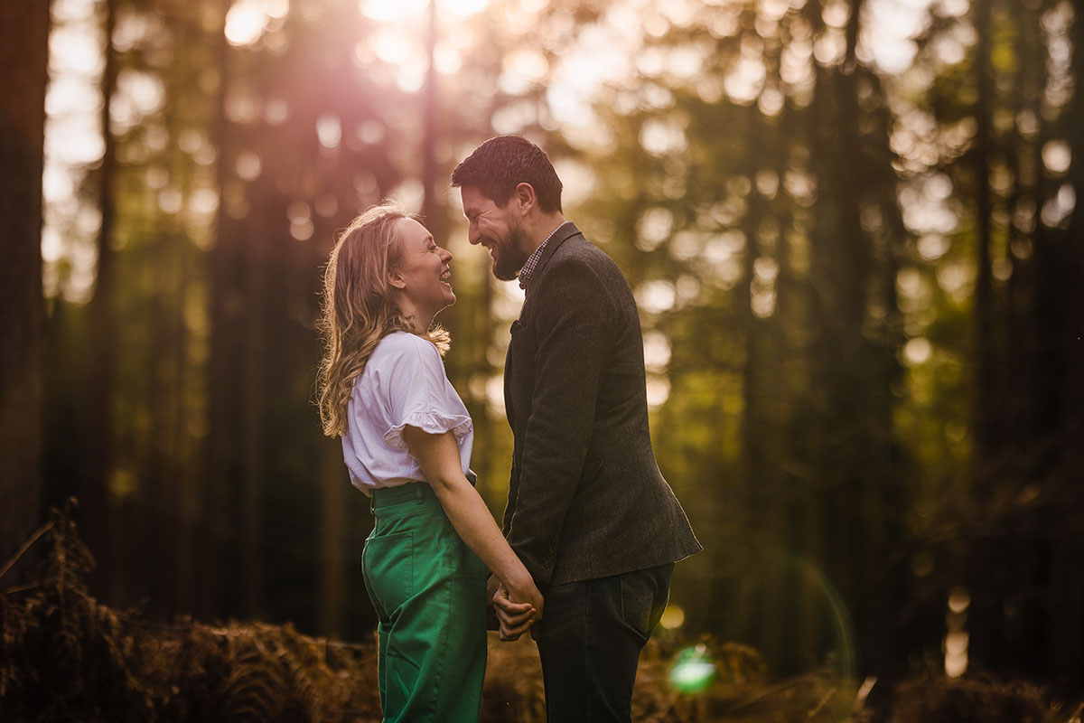 A heterosexual couple hold hands facing each other & laugh with woodland in the background