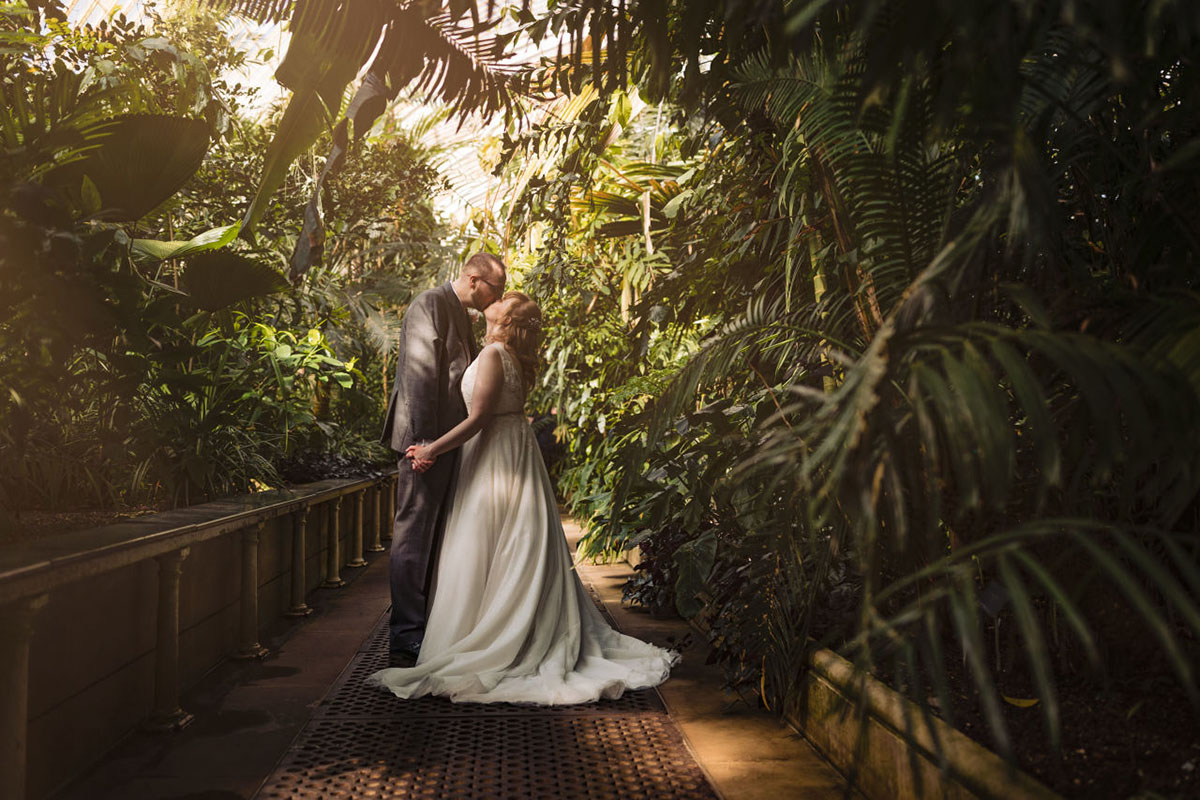 Bride & groom stand under tropical plants kissing in the Palm House