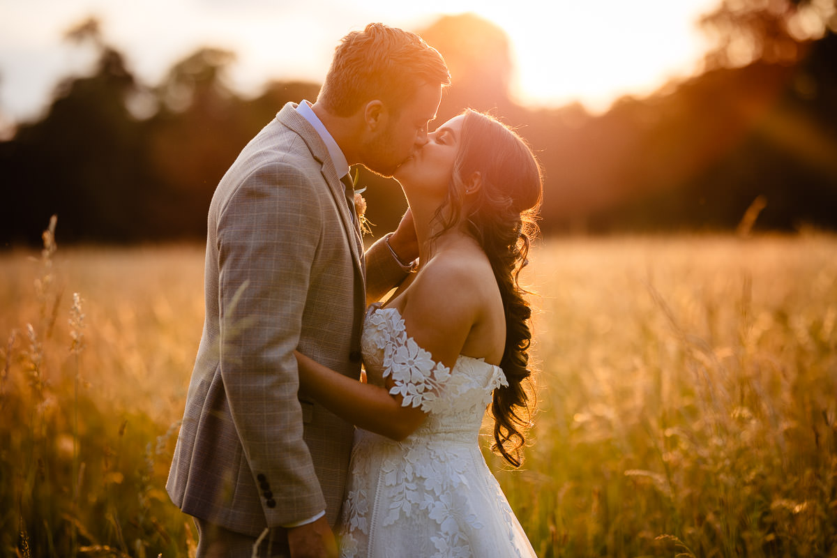 A bride & groom romantically kiss infront of the setting sun in the fields behind The Clock Barn