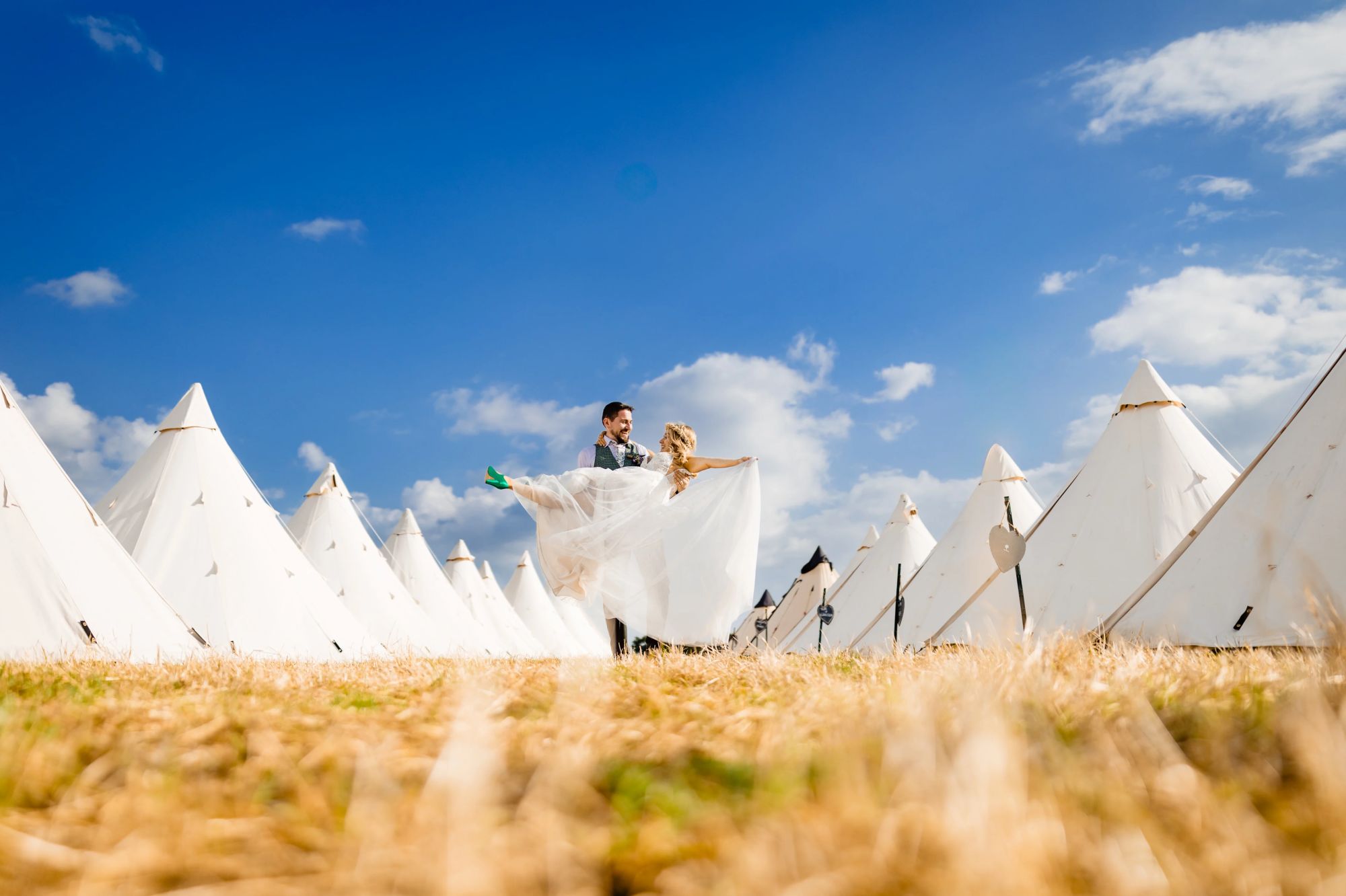 A groom holds his boho bride between two rows of Tipi's under a blue sky in a field of golden grass