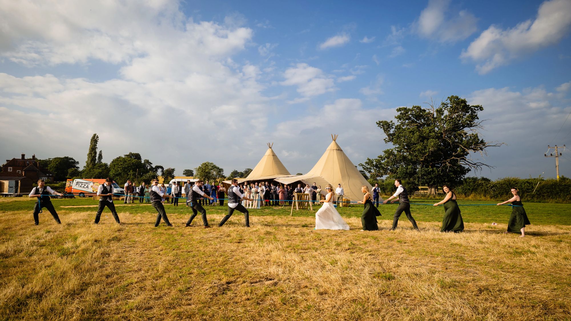 Wedding party in a tug of war located in a field in front of a Tipi under a blue sky