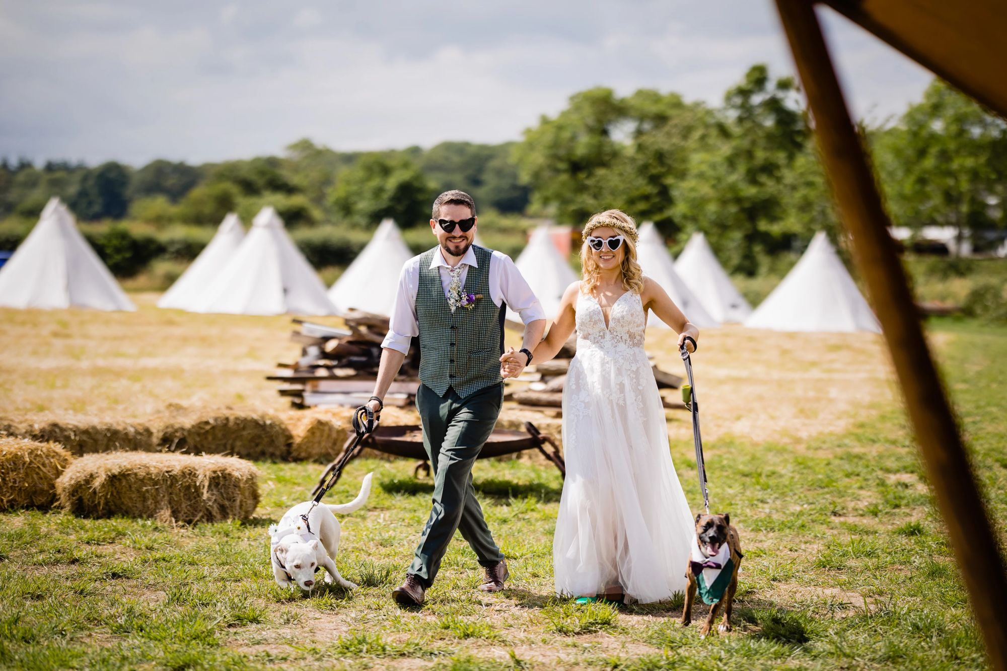 Boho styled bride & groom walk their dogs in a field in front of rows of white tents