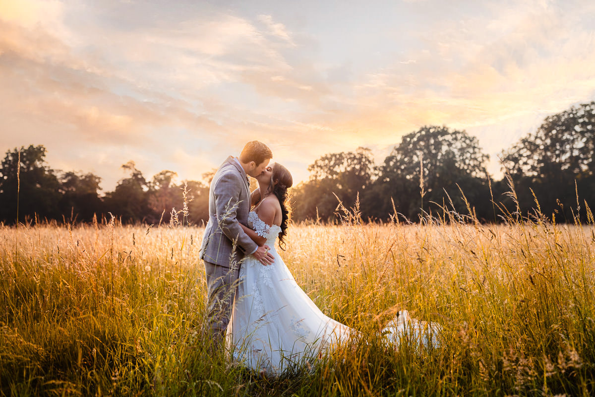 A portrait of a kissing bride & groom at sunset during golden hour in the field behind The Clock Barn