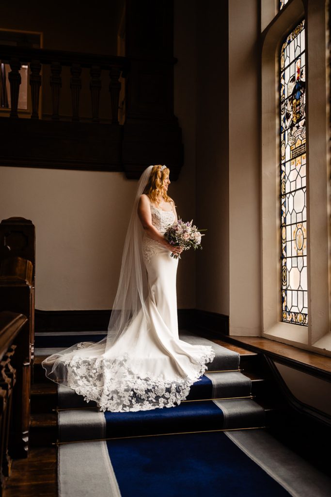 Full length portrait of a bride standing in front of the stained glass window at Easthampstead Park on her summer wedding day
