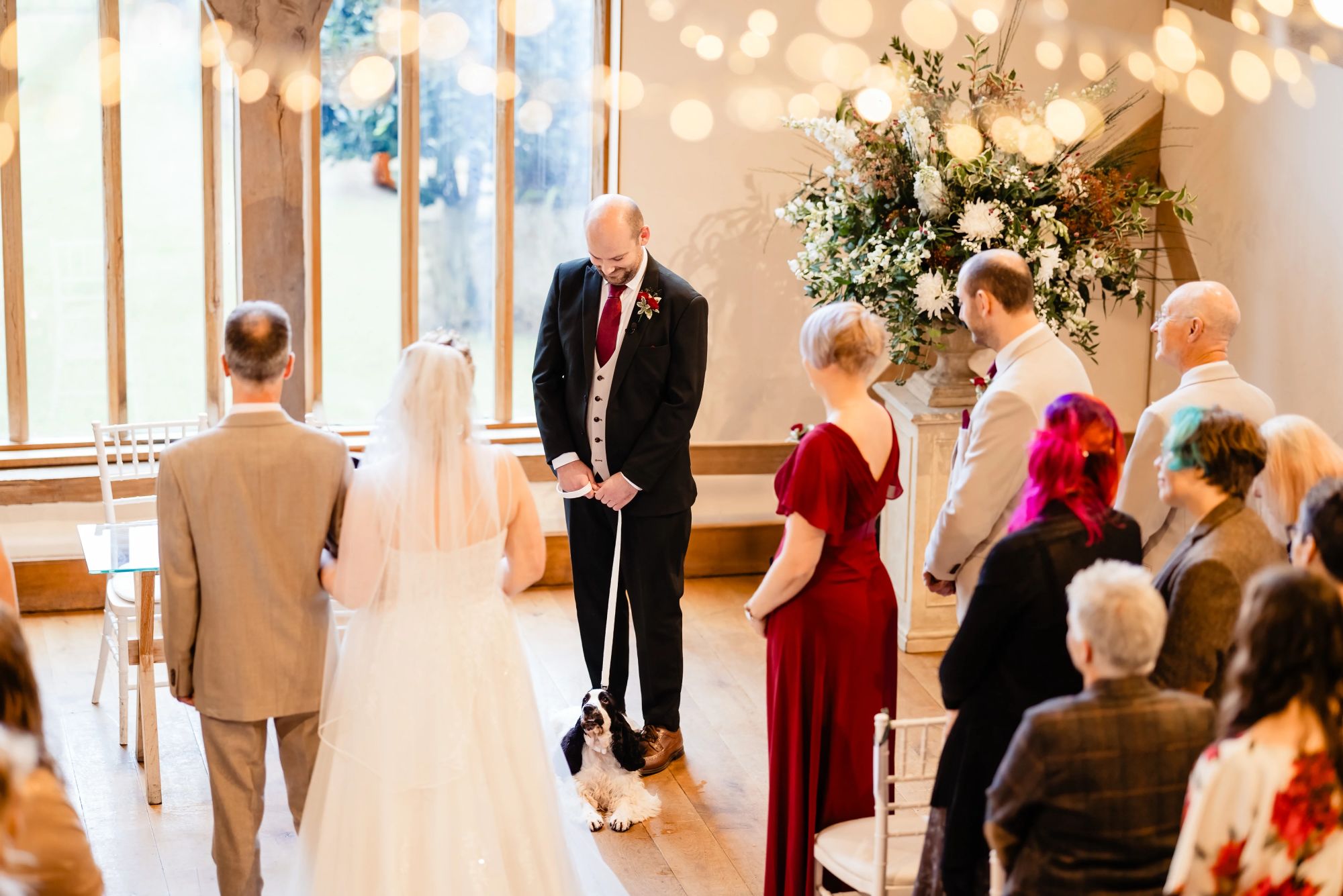Bride walking down the aisle at Cain Manor as groom waits with their spaniel dog sitting between them, surrounded by guests and fairy lights.